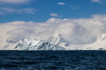Oceanside view of Antarctic Peninsula shoreline. Mountains with snow and ice, below a thick layer of clouds. Dark blue ocean in foreground; blue sky above. 
