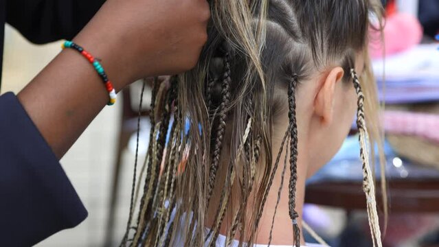 Young girl is sitting in a barber shop and waiting for a lot of pigtails to be braided on her head. Popular African pigtails are woven by a female African American master
