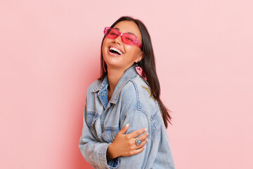 Smiling female posing isolated on pink background, wearing pink sunglasses and denim jacket, happy...