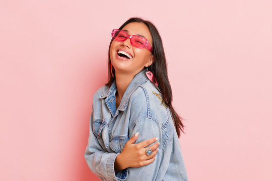 Smiling female posing isolated on pink background, wearing pink sunglasses and denim jacket, happy young woman holding herself with one hand, copy space, high quality photo