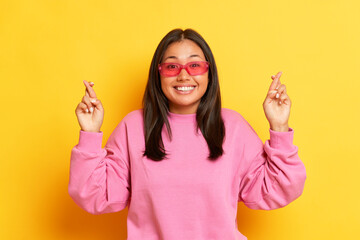 Pretty smiling girl in pink sunglasses standing on yellow background, student crosses her fingers for good luck before the exam, copy space, high quality photo