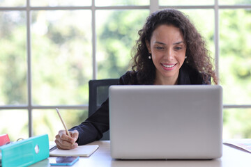 Attractive professional latin female employee worker sitting, using laptop computer with smart mobile phone at home workplace. Businesswoman working on paperwork while looking at camera and smile