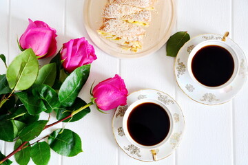 Beautiful festive morning breakfast. Pair of porcelain pattern cups of black coffee on saucer, fresh pastries sliced and bouquet of red roses on white background. Top view, flat lay