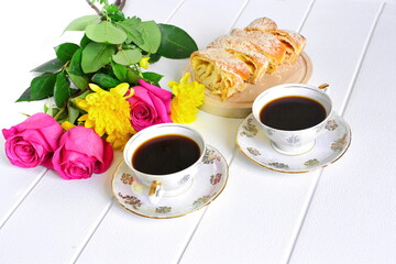 Beautiful festive morning breakfast. Pair of porcelain pattern cups of black coffee on saucer, fresh pastries sliced and bouquet of red roses and yellow chrysanthemums on white background. Copy space