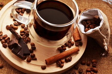 Glass cup of strong black hot aromatic coffee on wooden board between pieces of bitter chocolate, roasted coffee beans, cinnamon sticks and bag of grains on canvas