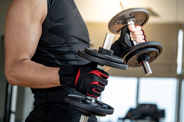 Sport man with well trained body in black sportswear wearing sport gloves lifting two dumbbells in fitness gym. Weight training and bodybuilding workout concept.