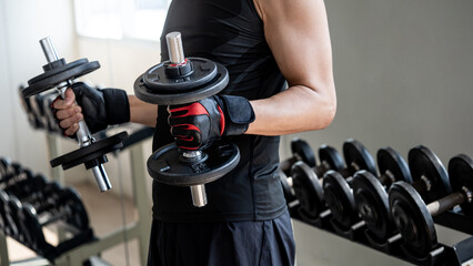 Sport man with well trained body in black sportswear wearing sport gloves lifting two dumbbells in...