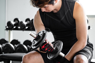 Sport man with well trained body in black sportswear wearing sport gloves sitting on workout bench lifting dumbbells in fitness gym. Weight training and bodybuilding concept.