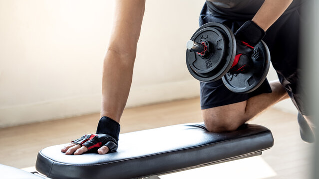 Sport man with well trained body in black sportswear doing one-arm dumbbell row on workout bench in fitness gym. Weight training and bodybuilding workout concept.