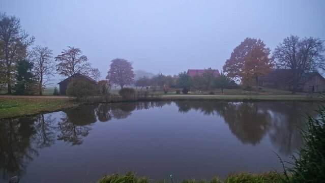 Timelapse of sunrise at lake on a green meadow with trees and wooden houses in cloudy and foggy day.
