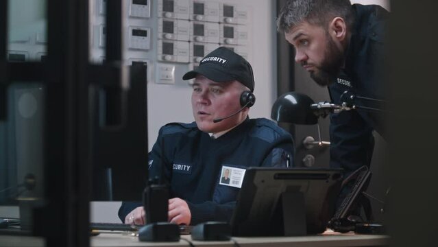 Man comes to colleague, looks view from security cameras displayed on computer and tablet monitors and talks by walkie talkie. Security officer sits at workplace and typing keyboard. CCTV technology.