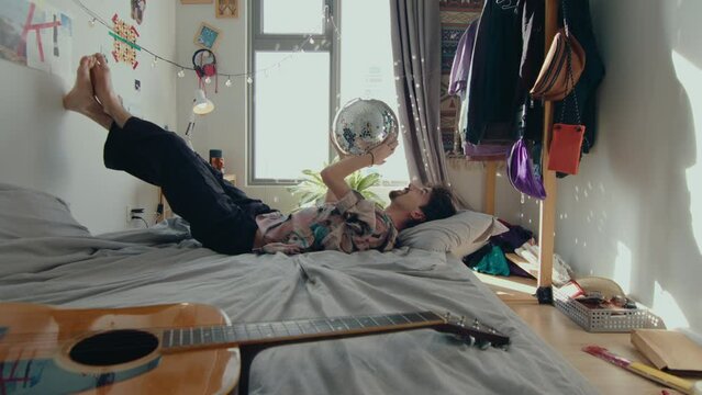 A Young Man Laying On A Bed With Foots On Wall With Disco Ball In Hands Reflecting Sun Rays Shining All Over The Room, Messy Tiny Room With Guitar And Big Window