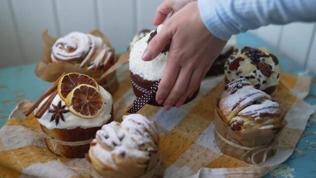 Traditional Easter cakes creatively decorated with white icing, candied fruits, nuts and chocolate, female hands take and lift one. Stylish authentic Easter buns on checked yellow tablecloth.