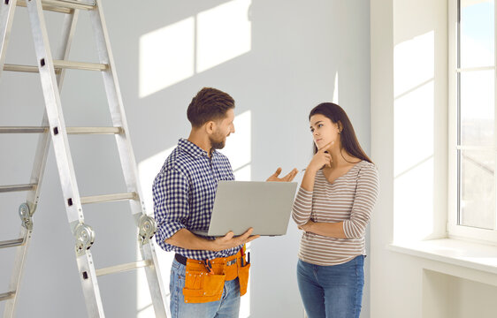 Repairman with modern laptop talking to young homeowner girl. Male worker with tool belt standing by step ladder, holding notebook computer and explaining to woman what he has to repair in the house