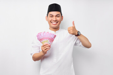 Smiling young Asian Muslim man showing thumbs up sign and holding cash money rupiah banknotes isolated on white background. People religious Islamic lifestyle concept