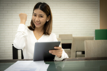 Beautiful asian office girl wearing white dress is sitting showing fist while holding tablet with smile on her face, Business woman cheering concept.