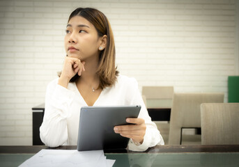 Beautiful asian business woman is sitting pretending to think about failed financial plan with a doubtful expression and holding tablet.