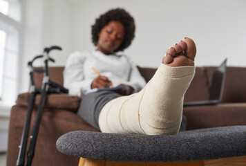 African American woman with a broken leg relaxing on the sofa at home, with her foot resting on a chair. Accident, injury, rehabilitation concept. Foot wrapped in bandage in closeup