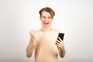 Fototapeta premium The guy uses a mobile phone, screams and shows a victory gesture on a white background.