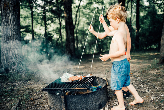 Brothers grilling hot dogs on barbeque grill while standing at campsite