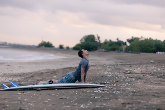 Side view of man performing cobra pose by surfboard at beach