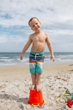Portrait of playful boy standing on bucket at beach against cloudy sky