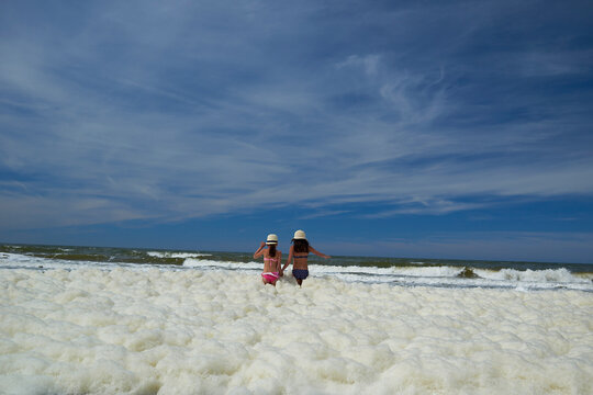 Rear view of sisters standing in sea foams while holding hands at beach against sky
