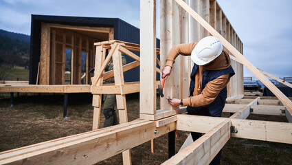 Man worker building wooden frame house on pile foundation. Carpenter hammering nail into wooden truss, using hammer. Carpentry concept.