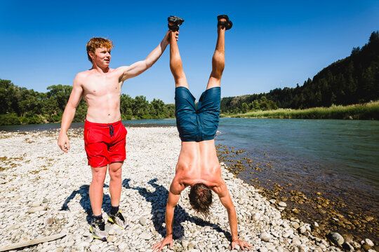 Teenage boy helping friend in doing handstand at lakeshore during summer