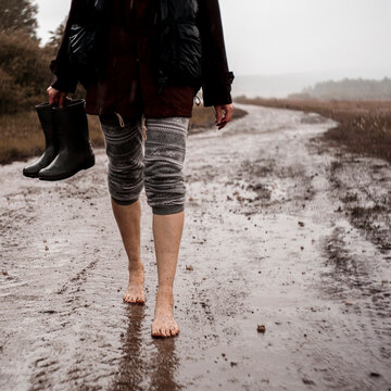 Low section of woman holding rubber boots while walking on wet dirt road