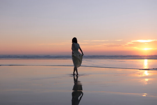 Carefree woman holding dress while walking at beach against sky during sunset