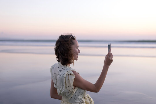 Side view of happy woman photographing with smart phone while standing at beach against sky