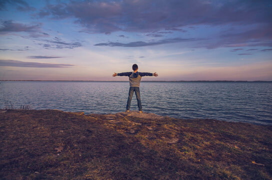Rear view of boy with arms outstretched standing against sea and cloudy sky during sunset