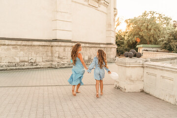 Daughter mother run holding hands. In blue dresses with flowing long hair, they hold balloons in their hands against the backdrop of a sunset and a white building.