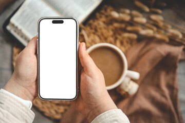 Hands holding a phone with an isolated screen, seen from above, with an open Bible, a coffee cup, and macarons in a home interior