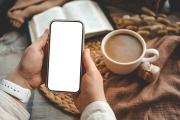 Hands holding a phone with an isolated screen, seen from above, with an open Bible, a coffee cup, and macarons in a home interior