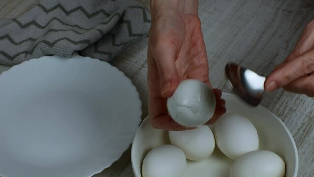 Cleaning a boiled chicken egg from the shell. Female hand breaks the shell of a boiled white egg with an iron tablespoon, puts the peeled shell in a plate, and puts the peeled chicken egg in bowl