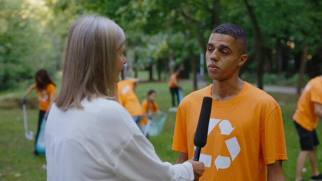 Female News Reporter Is Standing in Polluted Forest and Interviewing Multiethnic Eco Activist Who Are Collecting Trash. Group of Diverse Volunteers Cleaning Up the Public Park Background. News