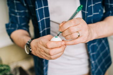 Easter day. Cropped hands of Adult man painting eggs on wooden background. Sitting in a kitchen with crayons. Preparing for Easter, creative homemade decoration.