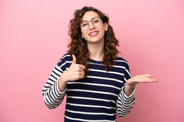 Young caucasian woman isolated on pink background holding copyspace imaginary on the palm to insert an ad and with thumbs up
