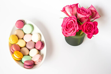 Colorful macaroons on a white ceramic plate and pink roses in a green vase on a white background.