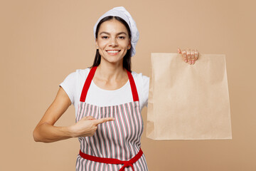 Young housewife housekeeper chef baker latin woman wears striped apron toque hat hold point on brown clear craft blank paper takeaway bag mock up isolated on plain beige background. Cook food concept.