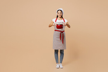 Fototapeta premium Full body smiling happy fun young housewife housekeeper chef baker latin woman wear apron toque hat hold in hand pot pan look camera isolated on plain pastel light beige background. Cook food concept.