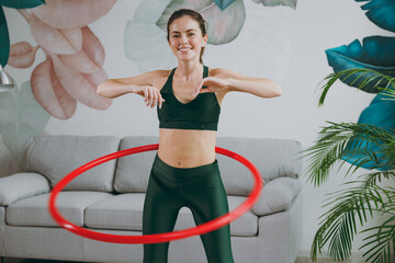 Young fun strong sporty athletic fitness trainer instructor woman wears green tracksuit hold using hula hoop for slim waist training do exercises at home gym indoor. Workout sport motivation concept - obrazy, fototapety, plakaty