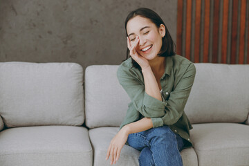 Obraz premium Young smiling shy happy woman of Asian ethnicity wears casual clothes look camera prop up chin sits on grey sofa couch stay at home hotel flat rest relax spend free spare time in living room indoor.