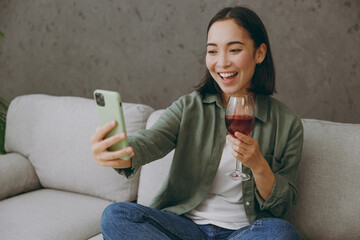 Young woman of Asian ethnicity wear casual clothes do selfie shot on mobile cell phone drink wine...
