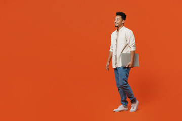 Naklejka premium Full body side profile view student young IT man of African American ethnicity wear light shirt casual clothes hold closed laptop pc computer walk go isolated on orange red background studio portrait.