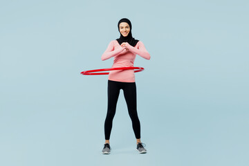 Full body young asian muslim fitness trainer sporty woman wear pink abaya hijab spend time in home gym use hula hoop look camera isolated on plain blue background studio Workout sport fit abs concept. © ViDi Studio