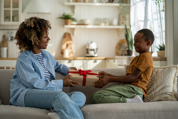 Happy African American woman mother receiving present from son while sitting together on sofa at home, holding gift box, child boy congratulating mom with mothers day or birthday, celebrate together