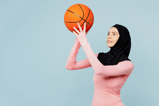 Young arabian asian muslim fitness trainer sporty woman wear pink abaya hijab spend time in home gym hold throw basketball ball isolated on plain blue background studio Workout sport fit abs concept. - Powered by Adobe